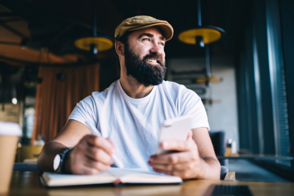 A man in a white t-shirt sitting at a coffee shop with a pen in one hand and his phone in the other as he is staring out of the window and smiling
