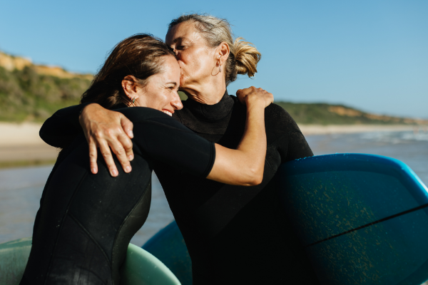An adult mother and daughter embracing on the beach while holding surf boards